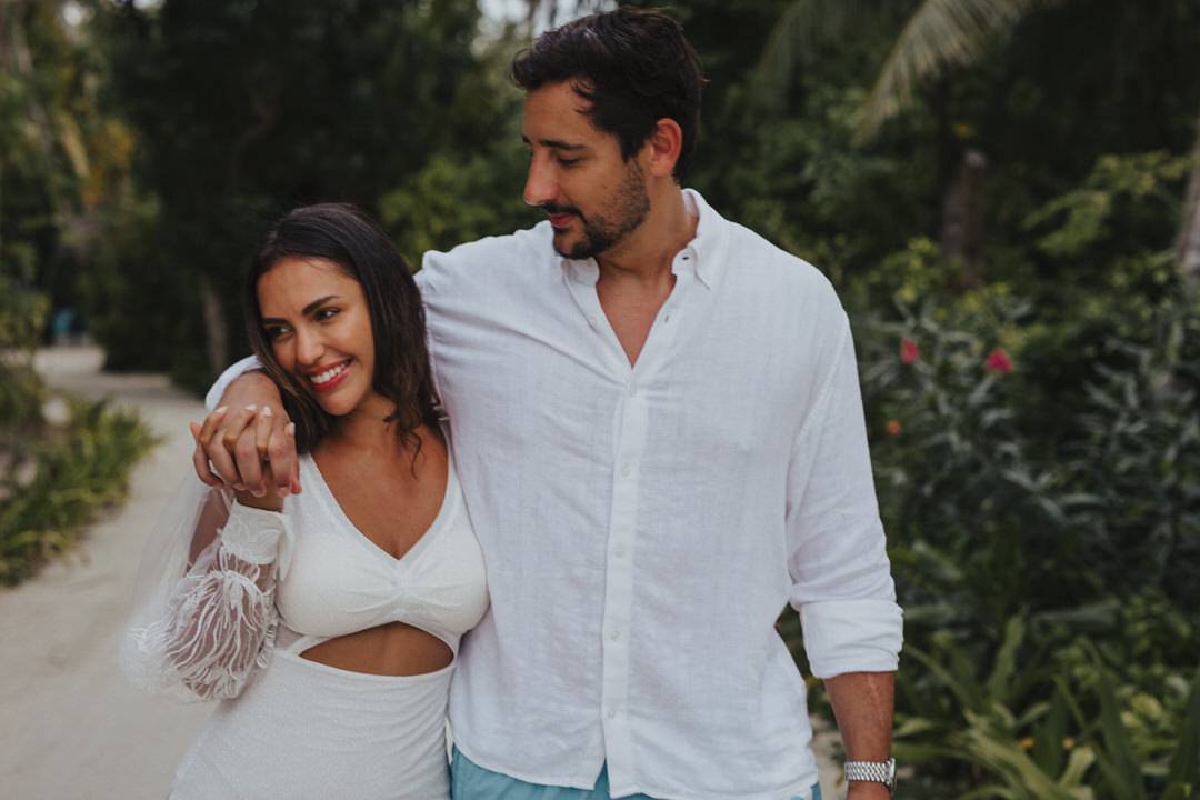 Brasilianisches Paar beim Spazierengehen durch den Sand auf den Malediven. Er umarmt sie über die Schultern. Beide lachen.